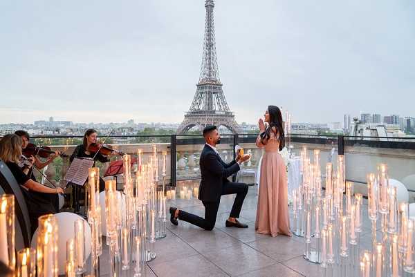 A marriage proposal is taking place on an outdoor rooftop terrace in Paris, with the Eiffel Tower visible directly behind the couple in the background. A man in a dark navy suit is down on one knee facing a woman in a blush pink floor-length gown, who has her hands raised to her face in reaction. The terrace is densely arranged with tall glass cylinder candle holders of varying heights, all lit, creating a warm candlelit atmosphere across the foreground and surrounding the couple. To the left, a string quartet of two violinists are performing, with sheet music stands in front of them. The scene is captured in a wide shot that balances the couple, the candle arrangement, the musicians, and the full Eiffel Tower in the composition.