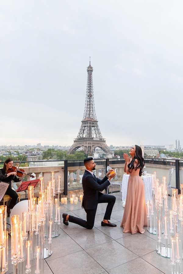 A marriage proposal taking place on an outdoor rooftop terrace in Paris, with the Eiffel Tower centered directly in the background. A man in a dark navy suit is down on one knee holding an open ring box toward a woman in a blush pink floor-length gown, whose hands are raised to her face in a surprised reaction. The terrace is decorated with dozens of tall glass candlestick holders of varying heights, all lit with candles, arranged across the tiled floor in a modern, romantic setup. A violinist dressed in black is seated to the left, playing from sheet music on a red music stand. A small draped table is visible to the right. The shot is a wide portrait-oriented image capturing the full scene including the city skyline.