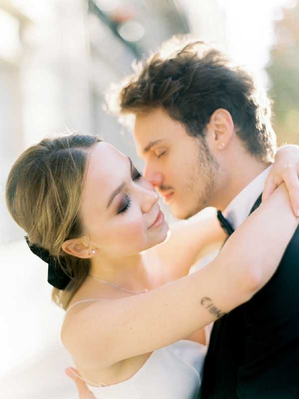 A close-up portrait of a bride and groom during what appears to be a couples portrait session. The bride wears a white strapless dress, has her hair pulled back with a large black bow, and wears pearl stud earrings; she has her arms wrapped around the groom's neck and is smiling softly with eyes closed. The groom wears a dark navy or black suit jacket with a black tie and leans in close to the bride's face. The bride has a small script tattoo visible on her forearm. The background is heavily blurred with warm, bright backlight creating a soft glow around the couple. The image has a film photography quality with warm, airy tones.