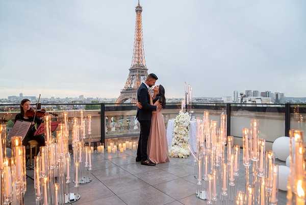 A couple shares an intimate moment on an open-air rooftop terrace in Paris, with the Eiffel Tower prominently visible in the background at dusk. The woman wears a dusty rose floor-length gown and the man is dressed in a dark navy suit, and they are embracing face-to-face in what appears to be a proposal or romantic portrait session. The terrace is styled with dozens of tall glass taper candle holders arranged across the floor, all lit, alongside a small round table adorned with a white floral arrangement of what appear to be roses and white blooms. To the left, a female violinist in dark clothing is seated and performing, adding a live music element to the setup. The overall decor palette is white and warm candlelight, creating a classic, intimate atmosphere. Wide-shot composition capturing the full scene and the iconic Parisian backdrop.
