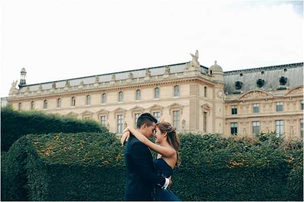 A couple shares an intimate embrace during what appears to be an engagement or pre-wedding portrait session in Paris, likely in the Tuileries Garden with the Louvre palace visible in the background. The man wears a navy blue suit and the woman wears a dark navy strapless dress, with her hair styled in a loose updo. The composition is a medium portrait shot with the couple centered against tall, neatly trimmed green hedges that frame them at shoulder height. The overall styling is classic and polished, with the ornate French classical architecture of the Louvre providing a grand architectural backdrop.