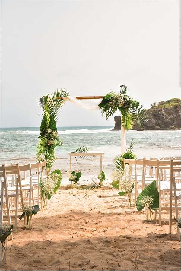 An outdoor beach wedding ceremony setup photographed before guests arrive, with the ocean and a rocky headland visible in the background. The ceremony aisle is lined with white folding chairs and aisle markers consisting of glass vases filled with baby's breath and large tropical leaves. A wooden arch frames the altar area, decorated with palm fronds, monstera leaves, white hydrangeas, baby's breath, and a draped length of white sheer fabric. A small white wooden ceremony table stands beneath the arch. The overall decor palette is white and green with a tropical theme. Wide shot capturing the full ceremony space from the back of the aisle.
