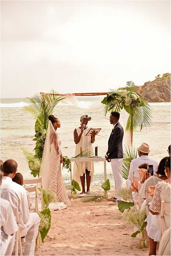 An outdoor beach wedding ceremony taking place directly on the sand with the ocean visible in the background. The bride wears a fitted white lace gown with a long veil and holds a tropical bouquet of large green leaves, while the groom wears a dark navy suit with white trousers; an officiant dressed in white stands between them holding a clipboard or book and speaking into a microphone. The ceremony arch is a bamboo or wooden frame draped with white sheer fabric and decorated with large tropical palm fronds, banana leaves, and clusters of white flowers, with matching floral arrangements of baby's breath in glass vases lining the aisle. Seated guests on white chairs, most dressed in white or light-colored attire, flank the aisle on both sides, with approximately 15-20 visible guests. The styling is tropical and relaxed with a natural green-and-white palette, and the wide-shot portrait composition captures the full ceremony setup with the coastline as a backdrop.