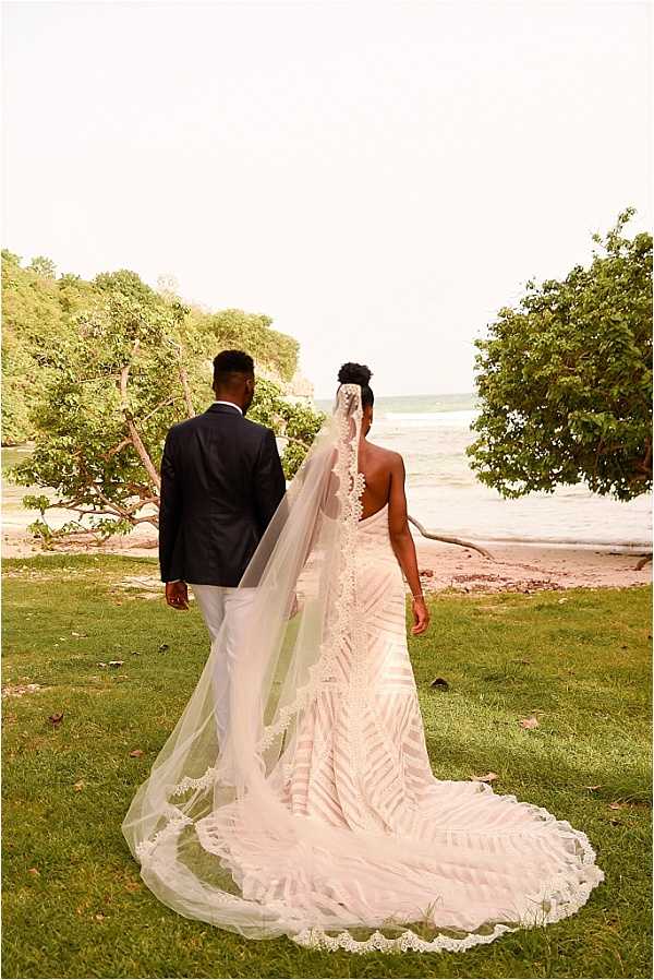 A couple portrait taken outdoors at a coastal beachside location, with the bride and groom photographed from behind as they walk toward the water. The groom wears a navy blazer with white trousers, while the bride wears a form-fitting backless gown with a bold leaf or geometric lace pattern and a cathedral-length veil trimmed with lace edging that trails across the grass. The bride's hair is styled in an updo. The composition is a full-length wide portrait shot capturing the full length of both the train and the veil spread across the lawn.
