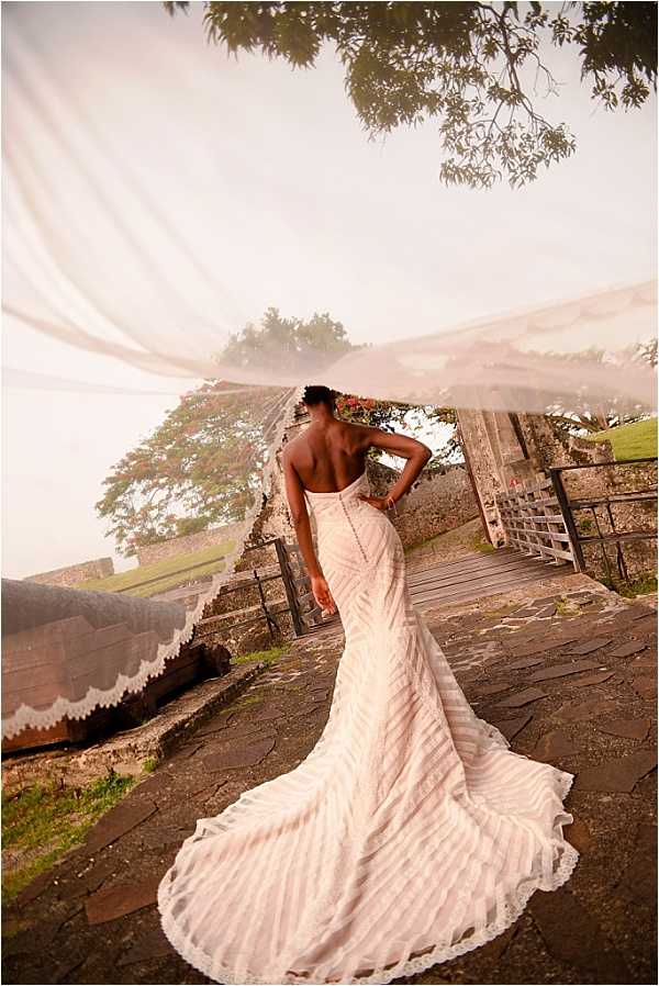 A bridal portrait taken outdoors at what appears to be a historic stone fortification or ruins, with wide stone steps, a wooden bridge or walkway, and low stone walls visible in the background. The bride stands with her back to the camera, wearing a fitted strapless ivory gown with horizontal stripe detailing, a low open back with button closures down the spine, and a long cathedral train spread across the stone ground. Her long cathedral-length veil with scalloped lace trim is caught in the wind and billows dramatically upward and across the frame, filling much of the upper portion of the image. The shot is a full-length portrait from behind, emphasizing the dress construction, the sweeping train, and the movement of the veil against the hazy, overcast sky.
