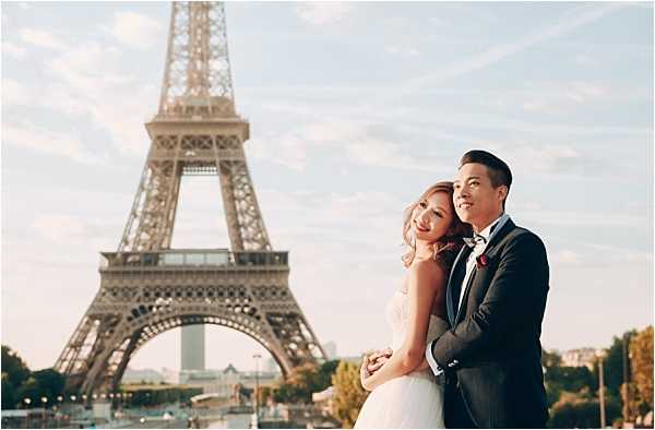 A couple poses for a portrait outdoors in Paris with the Eiffel Tower prominently centered in the background. The bride wears a strapless white gown and has her hair styled in loose waves, while the groom wears a navy suit with a white dress shirt and a red boutonniere. The groom stands behind the bride with his arms around her waist, and both are smiling. The shot is a mid-range portrait taken at ground level, with the Eiffel Tower softly out of focus behind them.