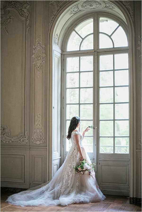 A bridal portrait taken indoors at what appears to be a French chateau, showing the bride standing in profile at a tall arched French door with white painted woodwork and a fanlight transom above. The bride faces away from the camera, her hand resting on the door handle, and holds a loose, garden-style bouquet featuring blush pink and white blooms with greenery. She wears an off-the-shoulder ball gown with heavy lace and floral appliqué detailing on a nude/ivory base, with a full tulle skirt and an extended cathedral-length train spread across the herringbone parquet floor. The room features ornate Rococo-style white plasterwork and raised wood paneling on the walls. The shot is a full-length portrait lit by natural light through the window, with soft, even tones. Potential venue feature image.