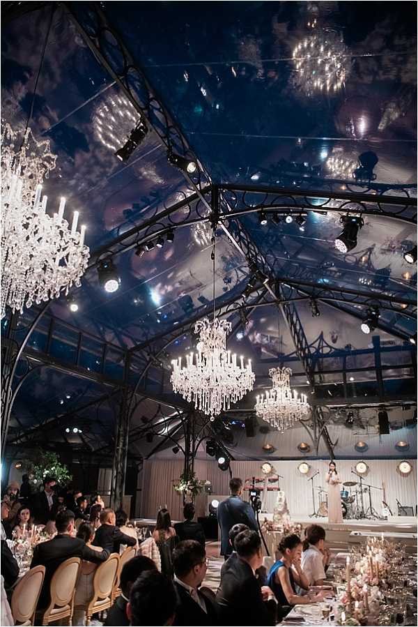 A wide-angle shot of a wedding reception dinner inside a large clear-roofed structure or conservatory-style tent, taken at night, allowing the dark sky to be visible through the transparent ceiling panels. Multiple large crystal chandeliers with candle-style lights hang from the black iron framework above the guests. Approximately 50 or more guests are seated at long banquet tables dressed with candles, glassware, and what appear to be blush-toned floral centerpieces. A live band and a speaker or performer are visible on a stage at the far end of the room, with professional stage lighting rigs mounted on the overhead structure. The overall decor palette combines crystal chandeliers, soft candlelight, and blush florals against the dark architectural framework, creating a classic formal reception atmosphere. The composition is a wide environmental shot taken from an elevated angle looking toward the stage.
