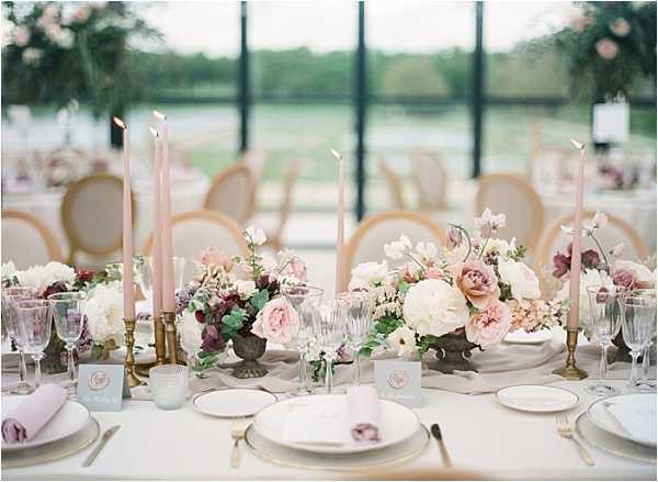 A close-up detail shot of a wedding reception tablescape set inside a glass-walled venue with a water view visible through the windows in the background. The table features multiple low floral centerpieces in antique patinated urns, composed of ivory peonies, blush garden roses, deep burgundy blooms, and greenery arranged along the center of the table. Tall blush pink taper candles in brass candlestick holders are placed between the arrangements. Place settings include white china plates with gold-rimmed chargers, crystal glassware, gold flatware, and folded blush pink linen napkins. Small grey place cards with circular printed details are positioned at each setting. The overall decor palette is blush pink, ivory, burgundy, and gold, styled in a romantic classic French aesthetic. Gold Louis-style chairs with cream upholstery are visible in the background.