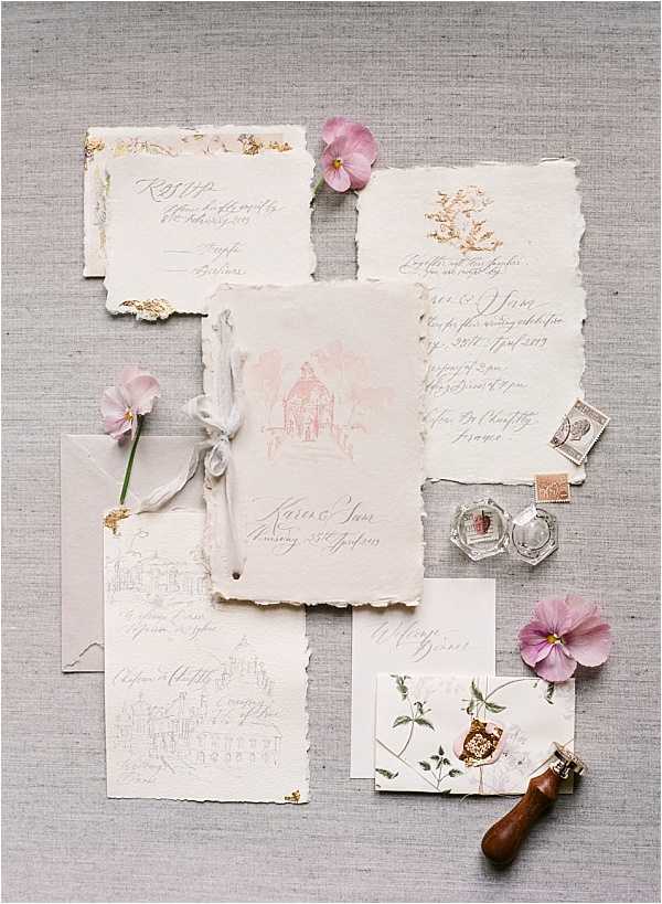 A flat lay detail shot of a wedding stationery suite arranged on a grey linen textile background. The suite includes multiple pieces of hand-torn deckle-edge paper in ivory and blush tones, featuring hand-calligraphed text, a watercolor illustration of a building on the main invitation, and gold leaf accents. Styling accessories include loose mauve and pink pansy and sweet pea blooms, a wax seal stamp with a wooden handle, two small glass ink wells, vintage postage stamps, and a small botanical-printed card with a red wax seal. The overall palette is blush, ivory, and gold with a romantic, handcrafted aesthetic.
