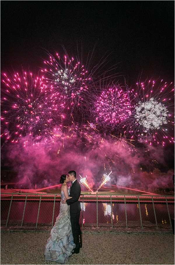 A couple shares a kiss during a fireworks display at night, standing in front of a metal railing with a reflective pool or fountain beyond it. The bride wears a layered, ruffled ball gown in light blue-grey tones with a strapless neckline, while the groom wears a dark suit with a white shirt and bow tie. The fireworks overhead burst in vivid magenta and white, filling the sky with dense pink smoke and trails of gold sparks, with the display reflected in the water behind them. The wide-angle portrait shot is taken at ground level, silhouetting the couple against the dramatic pyrotechnic backdrop in what appears to be a formal French garden or château grounds at night.