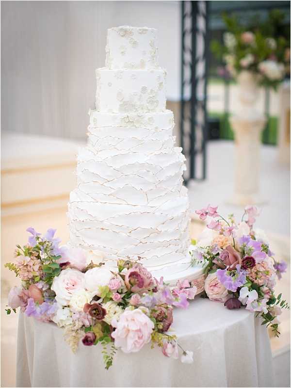 Close-up detail shot of a five-tier white wedding cake displayed on a round table covered with a pale grey linen. The cake features textured ruffled buttercream on the lower tiers and delicate white sugar floral appliqués on the upper tiers. The base of the cake is surrounded by a loose arrangement of blush garden roses, mauve ranunculus, lavender sweet peas, ivory hydrangea, and greenery. The overall floral palette combines blush pink, dusty mauve, and soft purple tones. The background is softly blurred, suggesting an indoor reception venue with large windows or open doors and a cream-toned urn visible in the background.