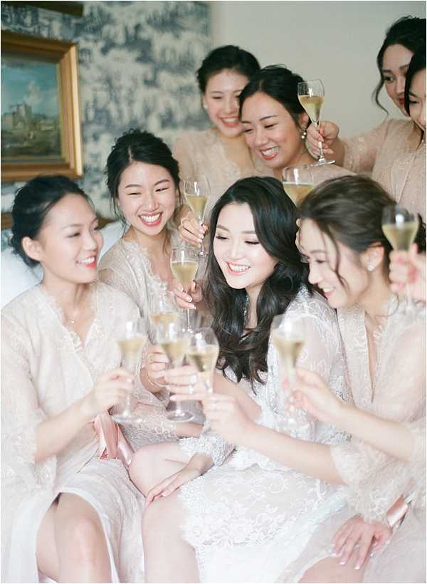 A getting-ready moment indoors showing a bride and approximately six bridesmaids toasting with champagne flutes. The bride, positioned at the center, wears a white lace robe and has her dark hair styled in loose waves, while the bridesmaids wear matching blush-toned lace robes. The group is seated closely together, laughing and smiling, in a room with blue-and-white toile wallpaper and a gold-framed painting visible in the background. The shot is a close-up portrait with soft, film-style tones and a slightly hazy quality consistent with fine art film photography.