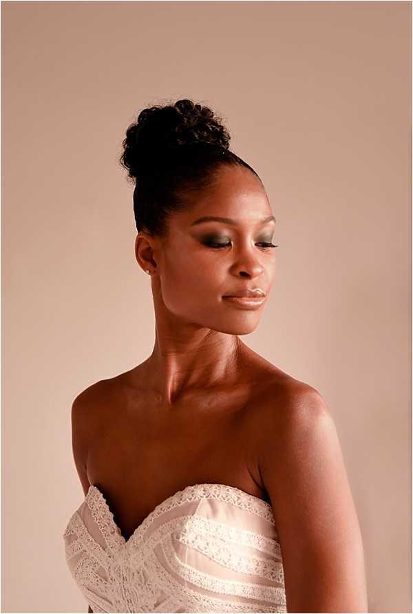 A close-up portrait of a bride shot against a warm beige studio backdrop. She wears a strapless ivory lace and satin corset-style bodice with scalloped lace trim along the sweetheart neckline. Her hair is styled in a high curly updo, and her makeup features smoky eye shadow with defined lashes and a neutral lip. She is looking downward with a calm expression. The lighting is soft and directional, creating subtle shadow definition across her collarbone and shoulders.