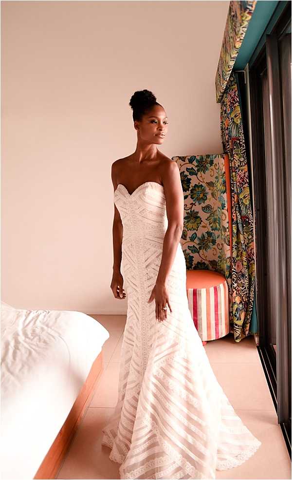 A bride stands alone in a hotel room or suite during the getting-ready phase, wearing a strapless ivory fit-and-flare wedding gown with geometric lace and bandage-style textured paneling and a flared train. Her hair is styled in a high bun and she wears a ring, gazing to the side. The room features boldly patterned floral fabric on a headboard or curtain panel in multicolor tones, a coral-orange round ottoman, and pink-and-white striped upholstery, giving the interior a colorful, eclectic feel. The shot is a full-length portrait taken indoors in natural light from a nearby window.