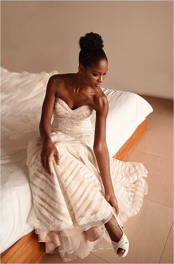 A getting-ready moment captured indoors, showing a bride seated on the edge of a low platform bed as she fastens her shoe. She is wearing a strapless sweetheart-neckline wedding gown in blush-ivory with tiered lace and tulle layers featuring scalloped trim, and a long cathedral-length veil draped beside her. Her hair is styled in a high updo bun. She is putting on a white strappy open-toe heel. The room has neutral beige walls and large-format tile flooring, giving a modern, minimal feel. The shot is taken from a slightly elevated angle, making it a portrait-style overhead composition.