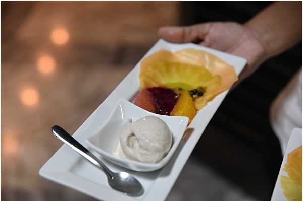 Close-up detail shot of a wedding reception dessert course being served by a staff member's hand. The white rectangular plate holds three components: a small square dish with a scoop of vanilla ice cream and a silver spoon, a portion of sliced citrus and blood orange segments, and a golden pastry cup filled with passion fruit. Warm bokeh lights are visible in the blurred background, suggesting candlelit reception decor. The presentation style is modern and plated, indicative of a formal sit-down dinner service.