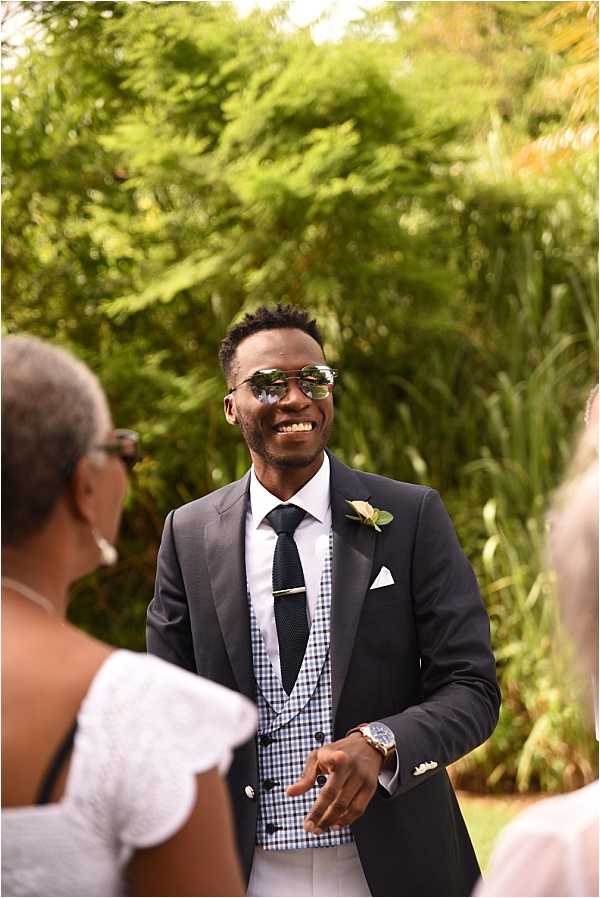 A groom stands outdoors during what appears to be a wedding ceremony, smiling broadly as he watches, likely seeing the bride approach. He is dressed in a dark navy blazer over a navy and white gingham check waistcoat, paired with a black knit tie secured with a tie bar, white dress shirt, and white trousers. He wears mirrored aviator sunglasses and a white boutonniere on his lapel, along with a statement wristwatch. Two guests are partially visible and out of focus in the foreground. The shot is a medium portrait taken from a slight angle, with a lush green garden backdrop.