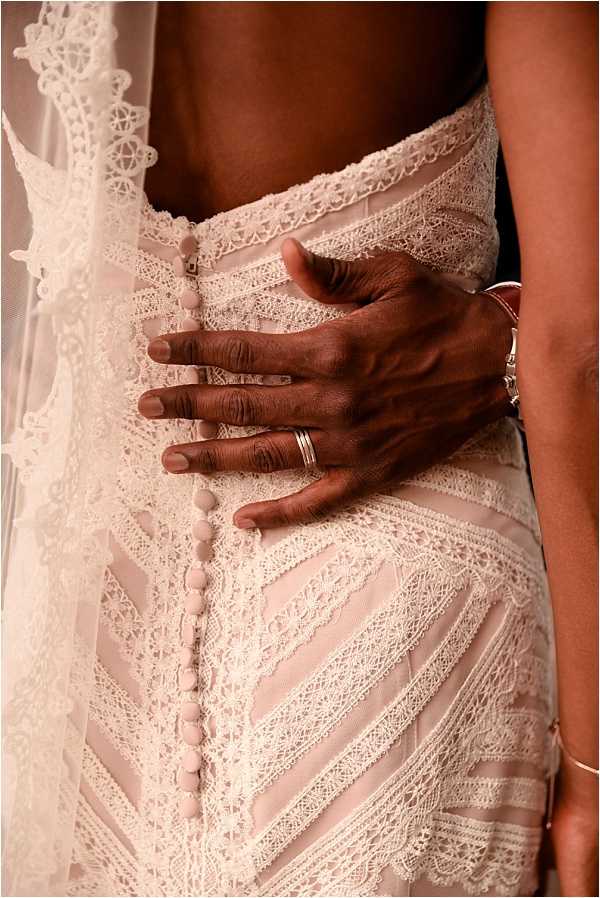 A close-up detail shot of a bride's back, showcasing an ivory lace wedding dress with a geometric chevron lace pattern and a column of fabric-covered buttons running down the spine. A lace-edged veil is visible along the left side of the frame. A hand — wearing silver stacked rings and a silver bangle bracelet — rests gently on the bride's waist, likely the groom's hand. The composition is tightly cropped to highlight the dress construction, accessories, and the contrast between the intricate ivory lace and the couple's skin tones.