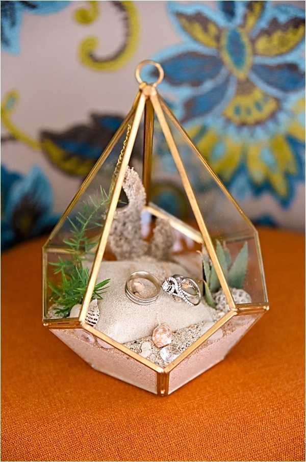 Close-up detail shot of two wedding rings — a plain silver band and a diamond engagement ring paired with a diamond wedding band — displayed inside a geometric gold-framed glass terrarium. The terrarium interior is styled with sand, small seashells, green fern sprigs, sage-colored succulents, and a small piece of dried coral, creating a coastal or beach-themed ring display. The terrarium sits on an orange fabric surface, with a blue and yellow floral embroidered textile visible in the soft-focus background.