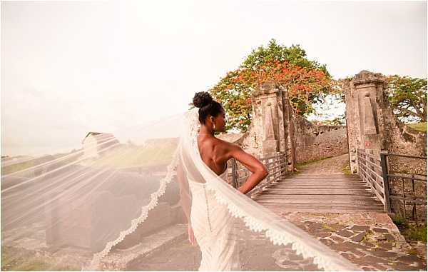 A bridal portrait of a solo bride standing on a wooden-railed stone bridge or walkway at what appears to be a historic fort or ruins. The bride wears a backless white gown with an upswept hairstyle and a long cathedral-length veil with lace trim that is caught in the wind and billows dramatically to the left of the frame. She is posed in three-quarter profile, looking away from the camera toward the ruins. A tree with red-orange blossoms is visible in the background between stone pillars. The image is a wide portrait shot with the flowing veil as a strong compositional element.