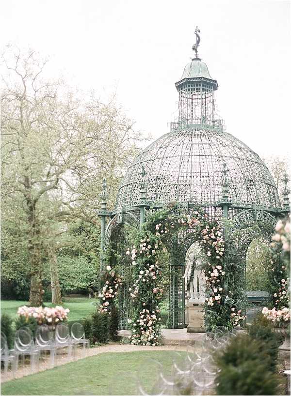An outdoor wedding ceremony setup in a formal garden, centered on a large ornate wrought-iron gazebo with a domed lattice roof and a weathered verdigris green patina, topped with a decorative finial. The gazebo entrance columns are wrapped in lush climbing greenery and blush pink and soft peach roses, with a classical stone statue visible inside the structure. Rows of clear acrylic ghost chairs line an aisle leading toward the gazebo, with low arrangements of blush and peach roses placed at the aisle ends. The composition is a wide shot taken from behind the guest seating, giving a full view of the ceremony structure. The overall decor palette is soft blush, peach, and green with a classic, romantic garden aesthetic. Potential venue feature image.