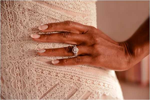 Close-up detail shot of a bride's hand resting against the bodice of her ivory lace wedding dress, showcasing a halo-style diamond engagement ring alongside a thin diamond wedding band on her ring finger. The nails are painted a soft blush-nude. The dress features intricate geometric lace paneling with layered crochet-style trim details. The image is tightly framed to highlight both the rings and the dress texture.