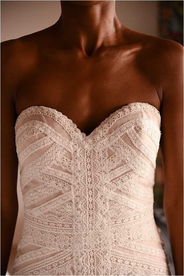 Close-up detail shot of a bride's wedding dress bodice during the getting-ready portion of the day. The dress features a strapless sweetheart neckline with intricate ivory lace appliqué arranged in a geometric, chevron-like pattern over a blush-pink satin underlayer. The lace detailing runs vertically and diagonally across the structured corset-style bodice, with delicate scalloped lace trim along the neckline edge. The image is cropped from the bride's collarbone to the waist, emphasizing the craftsmanship of the gown's construction.