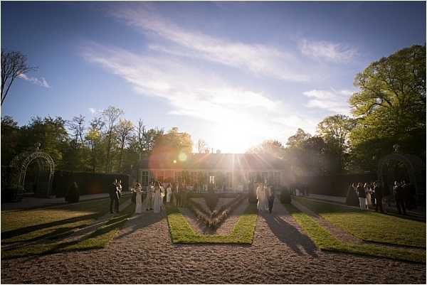 A wide-shot outdoor scene of a wedding cocktail hour or post-ceremony gathering on the formal grounds of a red-brick manor or château, taken at golden hour with strong sun flare and long shadows across the gravel and geometric grass parterre. Approximately 20–30 guests in formal attire are dispersed across the garden, with a group in white and light-colored dresses visible near the center-left, suggesting the bridal party. The venue features clipped hedges, a white metal arch or arbor on the left, and symmetrical lawn panels flanking a central gravel path leading to the building. The backlit composition creates dramatic lens flare and silhouette effects against the warm evening light. Potential venue feature image.