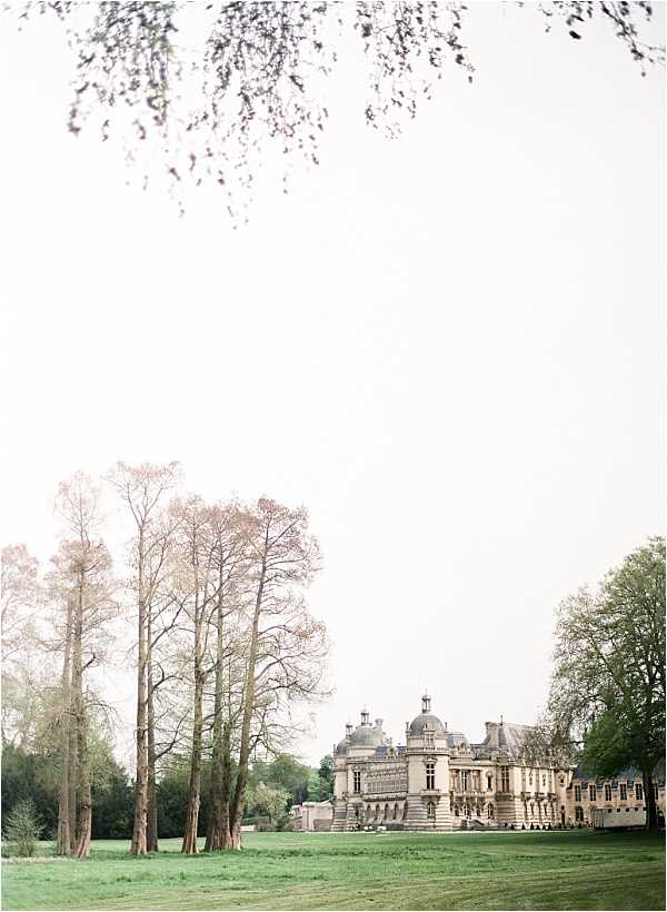 Wide-angle exterior shot of a grand French chateau with classical 17th-century architecture, featuring stone facades, dome-topped towers, and ornate detailing across multiple wings. The building is photographed from a distance across an open lawn, with tall bare trees framing the left side of the composition and leafy trees to the right. No people are visible in the frame. The image has a soft, airy, film-like quality with a pale, overexposed sky and muted tones throughout. Potential venue feature image.