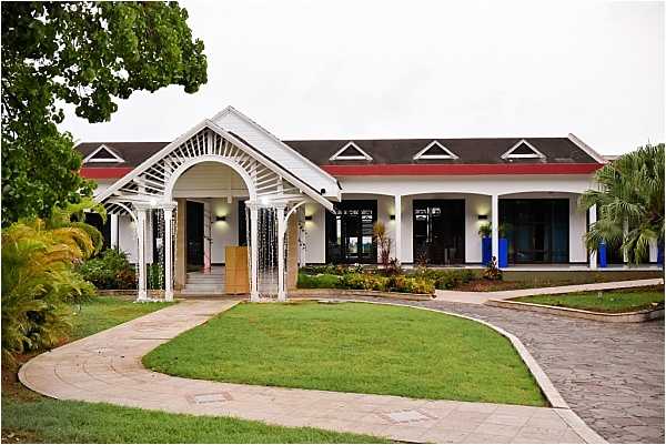 Wide exterior shot of a venue building featuring a white-painted single-story structure with a dark red roof, dormer windows, and a decorative white arched entrance pergola with hanging metallic curtain strands. The building has large dark-framed windows and wall-mounted lighting along its facade. The entrance is approached via a curved paved pathway surrounding a well-maintained lawn. No people are visible in the image. Potential venue feature image.