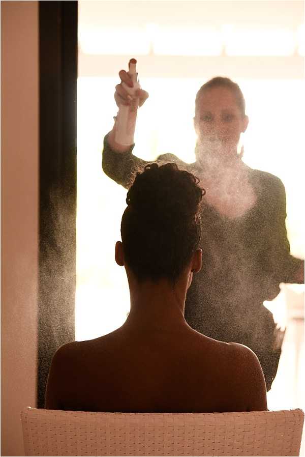 A getting-ready scene shot indoors, backlit by warm golden light from a large window. A hair stylist sprays hairspray over the bride's updo, creating a visible mist cloud between the two figures. The bride is seated in a white chair with her back to the camera, wearing no visible clothing on her upper back, with her dark hair styled into a curled updo. The composition is a medium portrait shot from behind, with both figures rendered largely in silhouette against the bright warm backlight, creating strong contrast between the dark foreground subjects and the glowing background.
