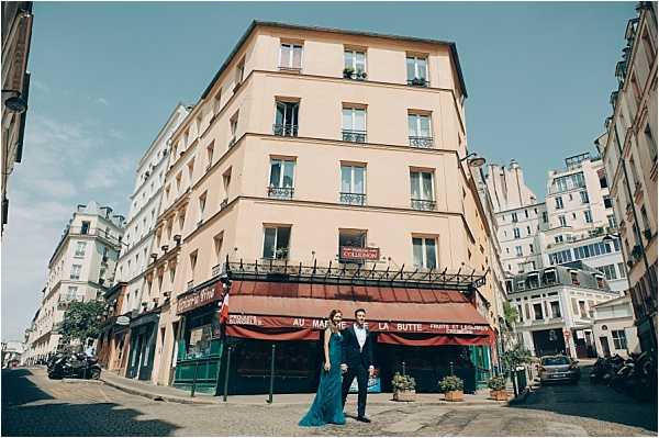 A couple poses together on a cobblestone street corner in Paris, likely in the Montmartre neighborhood, in front of a classic Haussmann-style building with a ground-floor shop bearing a red awning labeled 'Au Marché de la Butte.' The woman wears a floor-length teal/emerald gown and the man is dressed in a navy suit, both standing casually side by side. The shot is a wide environmental portrait capturing the full urban streetscape, including surrounding Parisian architecture, parked scooters, and street-level storefronts. The image has a relaxed, editorial feel consistent with a pre-wedding or post-wedding couple portrait session.
