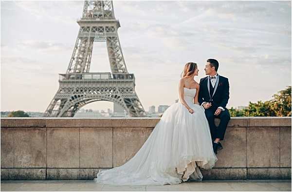 A couple poses for a portrait on the Trocadéro esplanade in Paris, with the Eiffel Tower prominently centered in the background. The bride wears a strapless white ballgown with a full tulle skirt and a long cathedral-length train that pools across the stone ledge, while the groom is dressed in a dark navy suit with a burgundy boutonniere and a black bow tie. The two are seated closely on a stone balustrade, turned toward each other in a relaxed, conversational pose. The shot is a medium-wide portrait taken at eye level, making the Eiffel Tower the dominant backdrop.