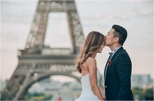 A couple poses for a portrait in front of the Eiffel Tower in Paris, shot outdoors with the iconic iron structure visible and softly out of focus in the background. The bride wears a strapless white fitted gown with her long hair worn loose and wavy, while the groom is dressed in a navy suit with a floral bow tie and a red pocket square, kissing her on the cheek. The composition is a medium close-up portrait framing both figures from roughly the waist up, with the Eiffel Tower centered between them. The overall styling is classic and polished, suggesting a pre-wedding or post-ceremony couple shoot at the Trocadéro.