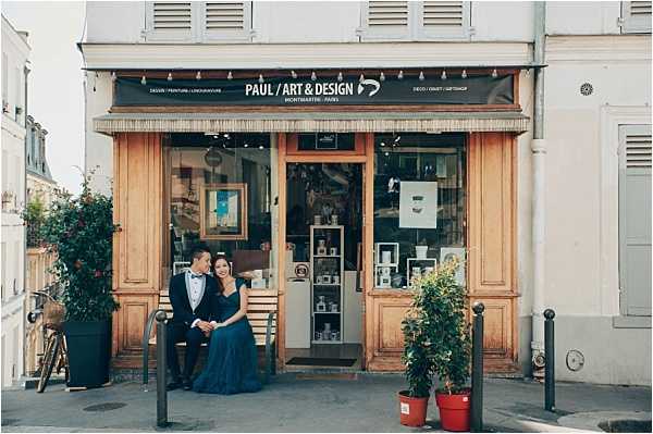 A couple poses seated on a bench in front of a Montmartre art and design shop called 'Paul / Art & Design' in Paris. The groom wears a navy suit with a black bow tie, and the bride wears a floor-length navy blue gown. The two are turned toward each other in a relaxed, intimate portrait. The shop front features warm wood paneling, display windows with artwork and decorative objects, and a dark signage banner. Wide shot composition capturing the full storefront and surrounding Parisian street context.