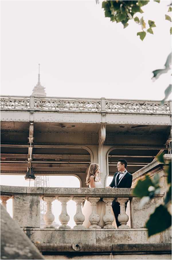 A couple poses for a portrait on the Bir-Hakeim bridge in Paris, with the Eiffel Tower visible in the background between the bridge's iron arches. The bride wears a strapless ivory or blush fitted gown and the groom is dressed in a dark navy or black tuxedo with a bow tie. The two are standing facing each other along a stone balustrade, framed by the ornate ironwork of the metro bridge above them. The shot is taken from a medium-wide angle at a slight distance, with tree branches framing the upper corners of the composition.
