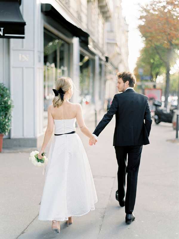 A couple walks hand-in-hand along a Parisian sidewalk in front of Haussmann-style building facades with black awnings, shot from behind in a full-length portrait. The bride wears a strapless, tea-length white dress with a black satin belt and a large black bow in her low blond ponytail, carrying a small bouquet of white flowers; the groom is dressed in a classic black suit with black dress shoes. The styling is minimal and graphic, built around a black-and-white color palette with the bride's hair accessory and belt as deliberate contrast elements. The image is composed as a mid-distance walking shot with soft warm daylight and a blurred street background.