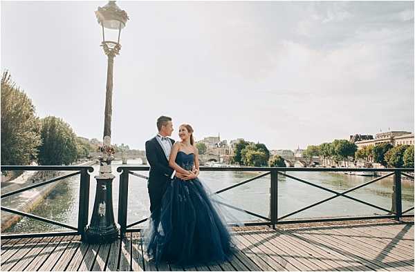 A couple poses for a portrait on a Parisian bridge over the Seine River, identifiable by the classic iron railing, wooden deck, and ornate lamp post characteristic of the Pont des Arts area. The bride wears a strapless navy blue tulle ballgown with a full skirt, and the groom is dressed in a dark navy suit with a white dress shirt and black bow tie. They stand facing each other in a relaxed embrace, with the Paris cityscape and river visible in the background. The shot is a wide portrait taken in natural daylight, capturing both the couple and the urban riverside setting.