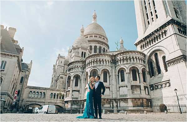 A couple poses together in front of the Sacré-Cœur Basilica in Montmartre, Paris, in what appears to be an engagement or pre-wedding portrait session. The woman wears a floor-length teal/peacock blue gown with a fitted silhouette and a small train, while the man is dressed in a dark navy suit with a bow tie. They stand close together on the cobblestone plaza in front of the basilica's rounded apse, with the white stone Romano-Byzantine architecture dominating the background. The shot is a wide environmental portrait that places the couple small against the grand scale of the building.
