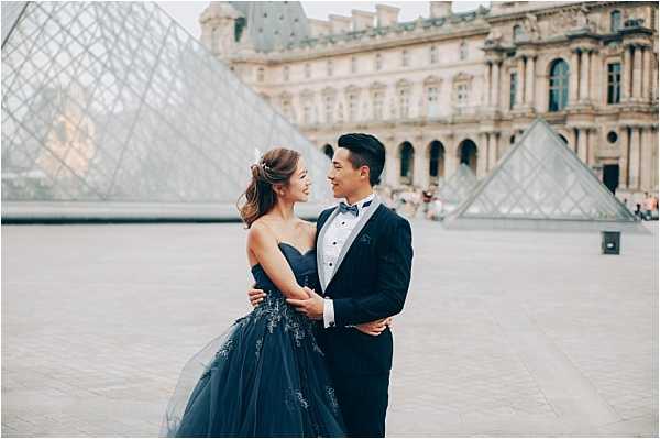A couple poses for a portrait shoot in the courtyard of the Louvre museum in Paris, with the iconic glass pyramid visible behind them along with the classical stone facade of the museum building. The bride wears a strapless navy blue ballgown with silver lace appliqué detailing and a full tulle skirt, paired with a half-up hairstyle and a small floral hair accessory. The groom is dressed in a navy blue tuxedo with a black bow tie and white dress shirt. The two face each other, holding hands and smiling, in a mid-distance portrait composition.