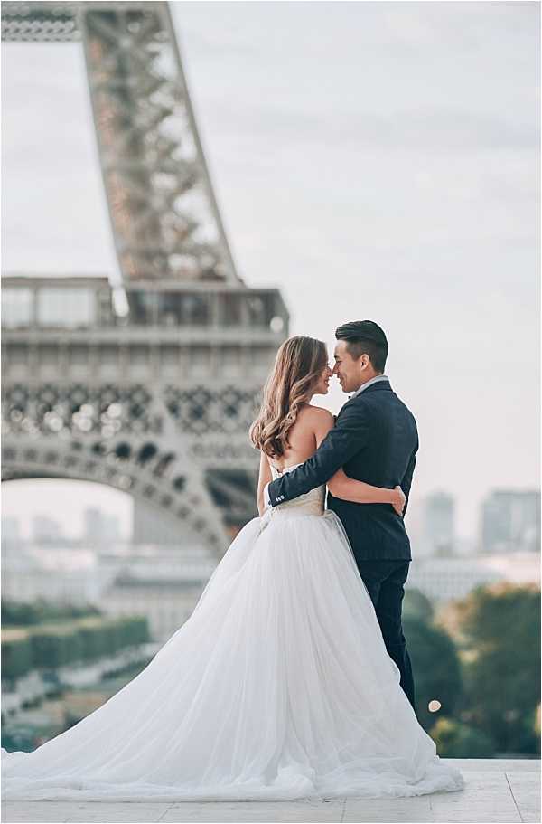 A couple poses for a wedding portrait outdoors in Paris with the Eiffel Tower prominently out of focus in the background. The bride wears a strapless ballgown with a voluminous white tulle skirt and a fitted bodice, her long wavy hair worn down, while the groom wears a dark navy suit. The two face each other with foreheads nearly touching in an intimate pose, with the groom's arms wrapped around the bride's waist. The shot is a mid-length portrait with shallow depth of field, placing full focus on the couple while the Eiffel Tower and Paris cityscape form a soft background.