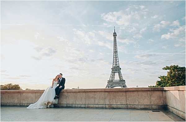 A bride and groom pose together on a wide stone terrace with the Eiffel Tower prominently visible in the background during golden hour. The bride wears a full tulle ballgown in ivory with a strapless bodice, and the groom is dressed in a dark navy suit; the couple is seated closely together on a low stone ledge. The shot is a wide portrait composition that emphasizes the Paris urban landscape and the scale of the Eiffel Tower relative to the couple. The styling is classic and formal, with soft warm light consistent with an early morning or late evening session.