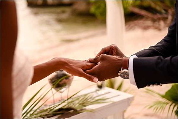 Close-up detail shot of a ring exchange moment during an outdoor beach ceremony, with the groom in a dark navy suit and white dress shirt placing a ring on the bride's finger. A small white ceremony altar or table is visible in the background, decorated with what appears to be a sand ceremony vessel and greenery. The setting is a sandy beach with tropical foliage and a body of water visible in the soft-focus background. The composition tightly frames the couple's hands, highlighting the ring and the groom's silver watch bracelet.