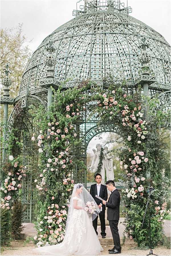An outdoor wedding ceremony taking place beneath a large ornate green wrought-iron domed gazebo structure, with the couple standing under a lush floral arch densely covered in blush and dusty rose garden roses interwoven with trailing greenery. The bride wears a white ball gown with lace appliqué detailing and a long cathedral veil, holding a bouquet of blush roses, while the groom is dressed in a black suit. An officiant stands between them holding papers, with a white stone angel statue visible through the arch in the background. The overall styling is romantic and classic with a garden-inspired palette of blush pink and green. Wide portrait shot capturing the full ceremony setup.