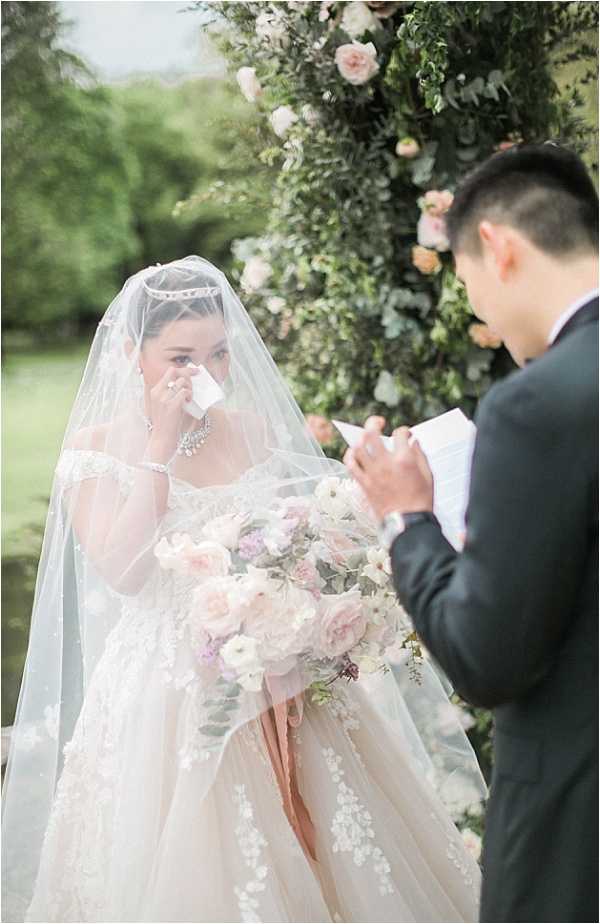 An outdoor wedding ceremony vow exchange between a bride and groom, set in front of a large circular floral arch decorated with blush and peach garden roses, white blooms, and trailing greenery. The bride, wearing an off-the-shoulder lace ball gown with floral appliqués, a long tulle veil, a crystal headpiece, and a diamond necklace, is visibly emotional and dabbing her eyes with a tissue while holding a large cascading bouquet of blush pink garden roses, white ranunculus, lavender blooms, and eucalyptus. The groom, in a dark navy suit, stands with his back partially to the camera reading from a small booklet of vows. The composition is a medium portrait shot focused on the bride's emotional expression, with the groom softly out of focus in the foreground right.