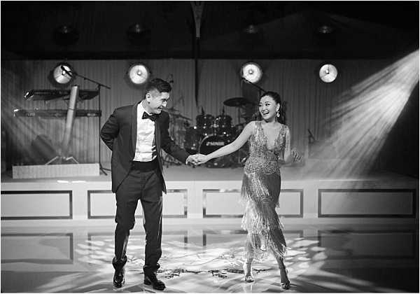 Black-and-white wide shot of a couple sharing their first dance on a polished dance floor during an indoor wedding reception. The groom wears a classic black tuxedo with bow tie, and the bride wears a fringed, heavily embellished midi-length dress with a deep V-neckline, consistent with a 1920s Art Deco or Great Gatsby-inspired styling theme. They are holding hands and smiling at each other, caught mid-movement. Behind them, a live band stage is visible with a drum kit and keyboard setup, and four stage spotlights beam dramatically downward, creating strong contrast and visible light rays across the scene. The high contrast black-and-white tones emphasize the theatrical lighting and the texture of the bride's fringed dress.