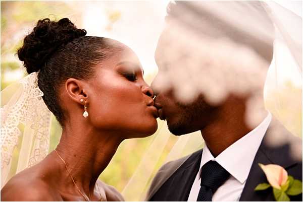 Close-up portrait of a bride and groom sharing a kiss outdoors, partially framed by the bride's lace-edged ivory veil draped across the foreground. The bride wears pearl drop earrings, a delicate gold necklace, and has her hair styled in an updo with natural texture; her dress appears to be a strapless style in a light tone. The groom wears a dark charcoal suit with a white dress shirt and a dark navy tie, and has a peach-toned flower boutonniere on his lapel. The shot is a tight couple portrait with a shallow depth of field, with the veil creating a soft blur effect over part of the groom's face.