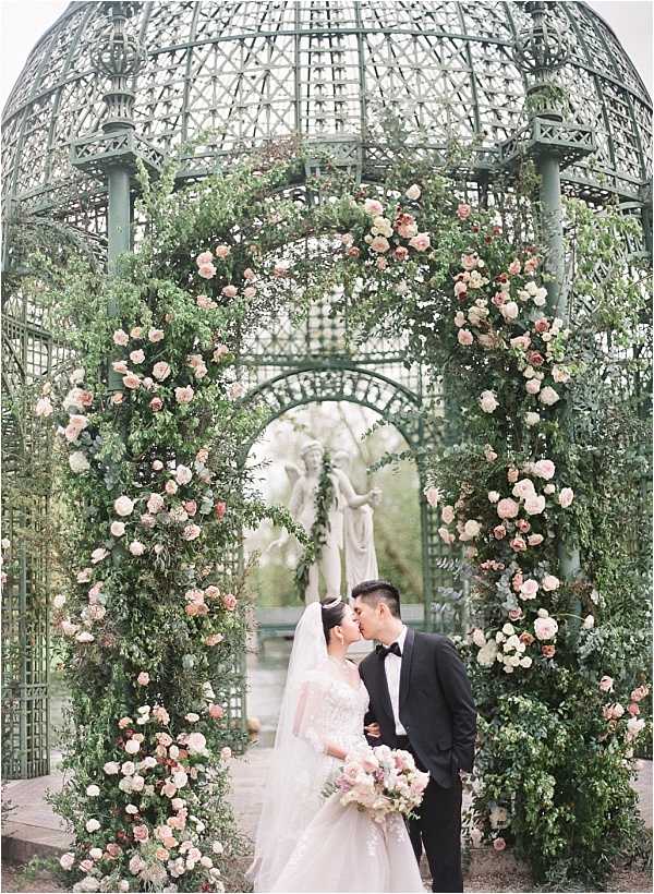 A bride and groom share a kiss during an outdoor couple portrait session beneath a large ornate green wrought-iron gazebo with a domed lattice roof. The gazebo arch framing the couple is heavily decorated with lush greenery and blush pink, dusty rose, and ivory garden roses interspersed throughout. The bride wears a long-sleeved lace wedding gown with a cathedral veil and holds a loose bouquet of blush and ivory roses with eucalyptus, while the groom wears a black tuxedo with a black bow tie. A classical white stone figurine sculpture is visible in the background at the center of the gazebo. The overall styling is romantic and garden-inspired with a soft blush and green floral palette. This is a medium portrait shot with the architectural structure used as a dramatic frame around the couple. Potential venue feature image.