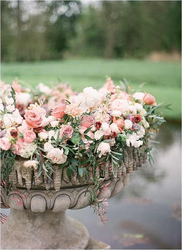 A close-up detail shot of a large floral arrangement displayed in a weathered stone urn pedestal, positioned outdoors near a reflective pond or water feature. The arrangement is densely packed with blush pink garden roses, peach roses, ivory peonies, dusty mauve ranunculus, deep burgundy anemones, cream sweet peas, and trailing olive-toned greenery and eucalyptus that cascades over the sides of the urn. The overall floral palette is a mix of blush, peach, cream, and deep burgundy tones, creating a lush, overflowing garden-style arrangement consistent with a romantic classic aesthetic. The background is softly out of focus, revealing a manicured lawn and treeline.